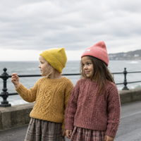 Two young girls standing on a seaside promenade under an overcast sky. The girl on the left wears a mustard yellow cable-knit sweater and matching beanie, while the girl on the right wears a dusty rose knit sweater and a pink beanie.