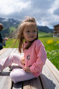 A young girl with long light brown hair sits on a wooden bench outdoors, wearing a pink long-sleeve top with a small black cat graphic and matching pink leggings. The background shows a blurred mountain landscape, green meadow, and cloudy sky.