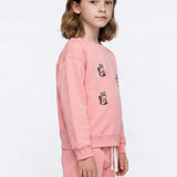 A young girl with shoulder-length brown hair stands in profile wearing a light pink sweatshirt and matching sweatpants. The sweatshirt features small black and white cat illustrations on the chest, and the pants have a white drawstring.