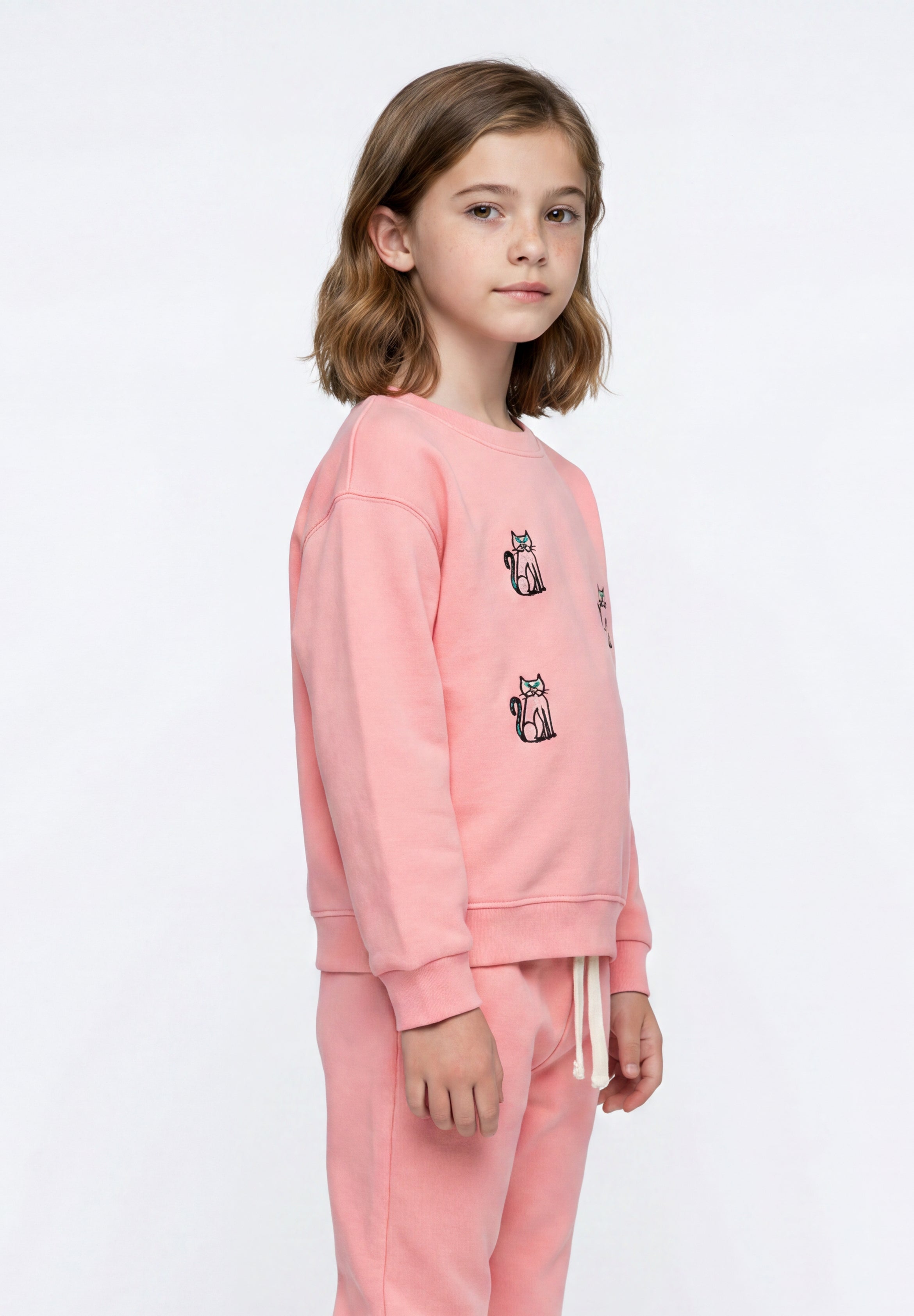 A young girl with shoulder-length brown hair stands in profile wearing a light pink sweatshirt and matching sweatpants. The sweatshirt features small black and white cat illustrations on the chest, and the pants have a white drawstring.