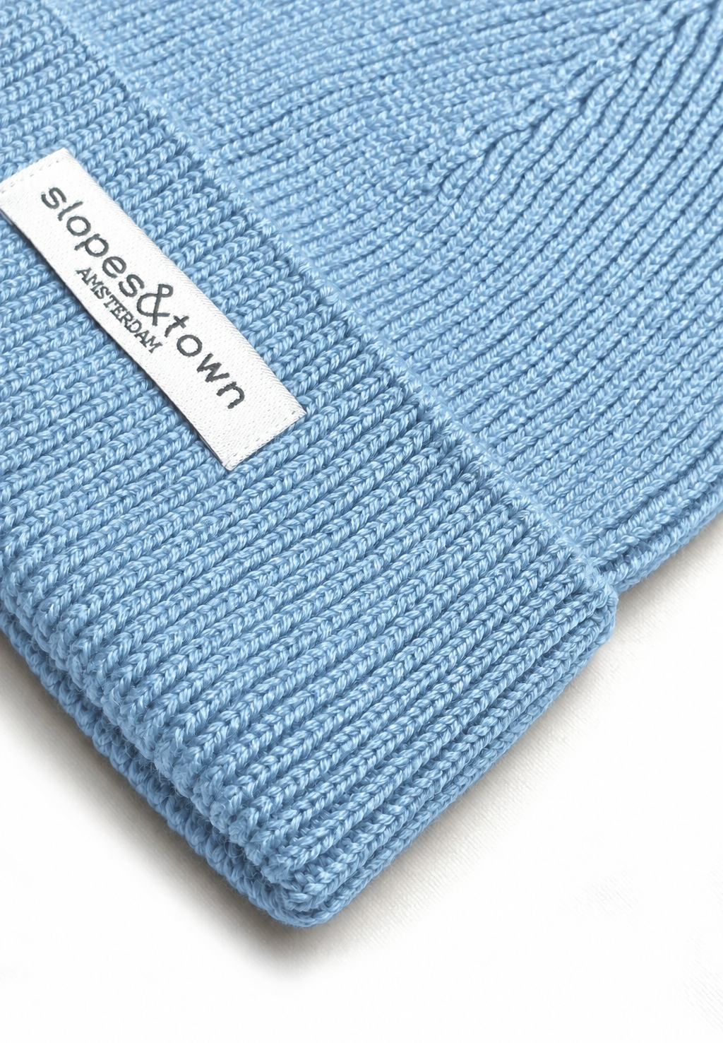 A close-up of a light blue ribbed knit beanie with a white fabric brand label reading 'slopes&town Amsterdam' on the cuff.