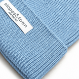 A close-up of a light blue ribbed knit beanie with a white fabric brand label reading 'slopes&town Amsterdam' on the cuff.