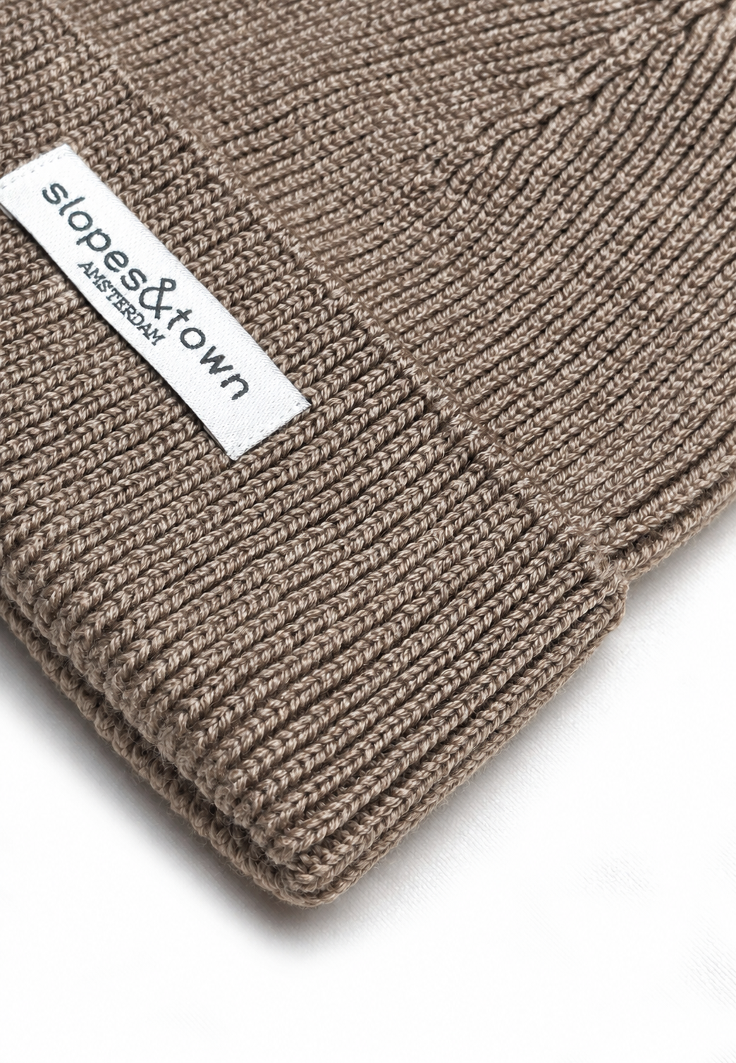 Close-up of a ribbed merino wool knit beanie in taupe brown with a white fabric brand label reading 'slopes&town AMSTERDAM'.