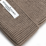 Close-up of a ribbed merino wool knit beanie in taupe brown with a white fabric brand label reading 'slopes&town AMSTERDAM'.