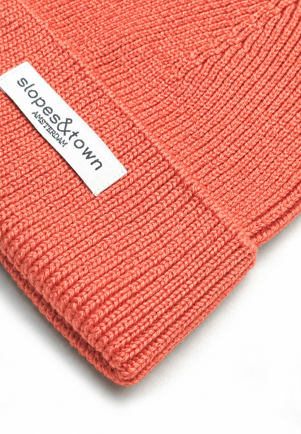 Close-up of a coral pink ribbed knit beanie with a white fabric label reading 'slopes&town AMSTERDAM'.