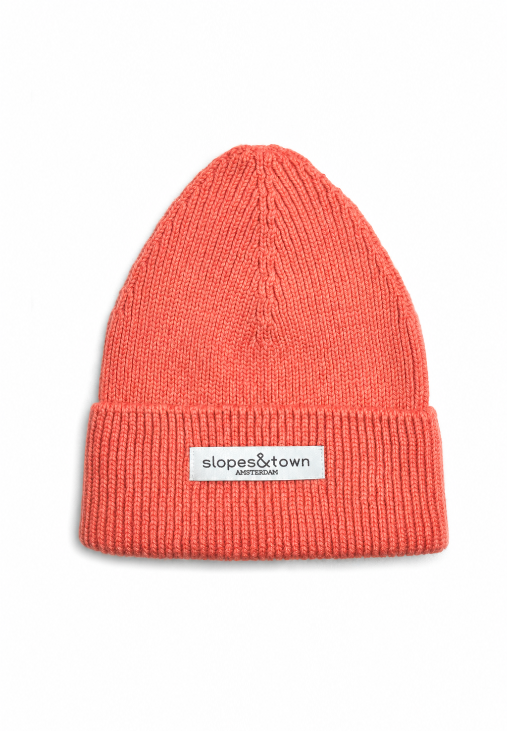 A coral pink ribbed knit merino wool beanie hat with a white rectangular brand patch on the front cuff that reads 'slopes&town Amsterdam. 