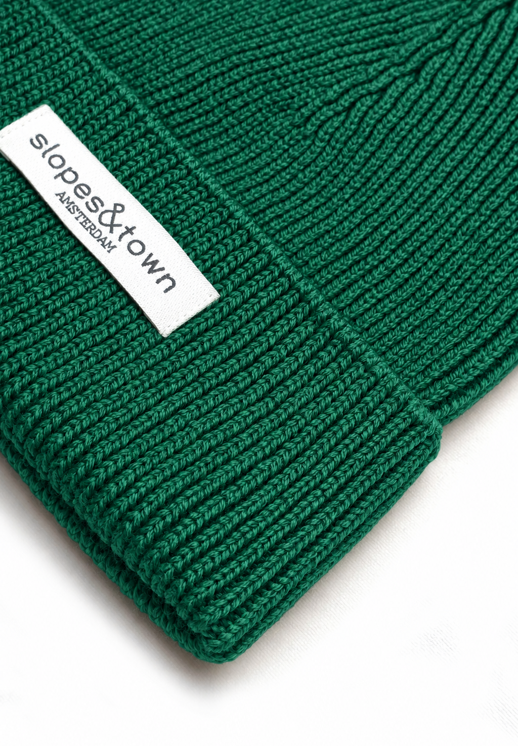 A close-up of a dark green ribbed knit beanie with a white fabric brand label reading 'slopes&town amsterdam'.