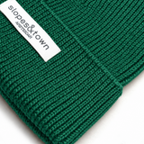 A close-up of a dark green ribbed knit beanie with a white fabric brand label reading 'slopes&town amsterdam'.
