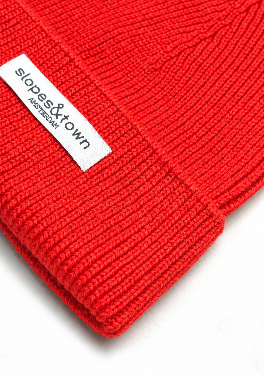 A close-up of a bright red ribbed knit beanie with a white fabric label reading 'slopes&town AMSTERDAM' on a white background.