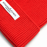 A close-up of a bright red ribbed knit beanie with a white fabric label reading 'slopes&town AMSTERDAM' on a white background.