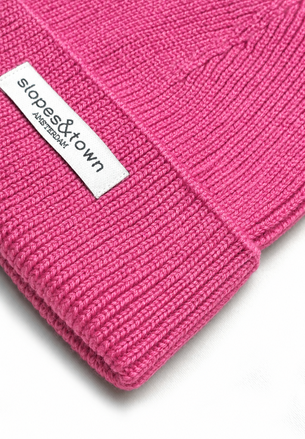A close-up of a bright pink ribbed knit beanie with a white fabric brand label reading 'slopes&town AMSTERDAM' on the cuff