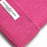 A close-up of a bright pink ribbed knit beanie with a white fabric brand label reading 'slopes&town AMSTERDAM' on the cuff