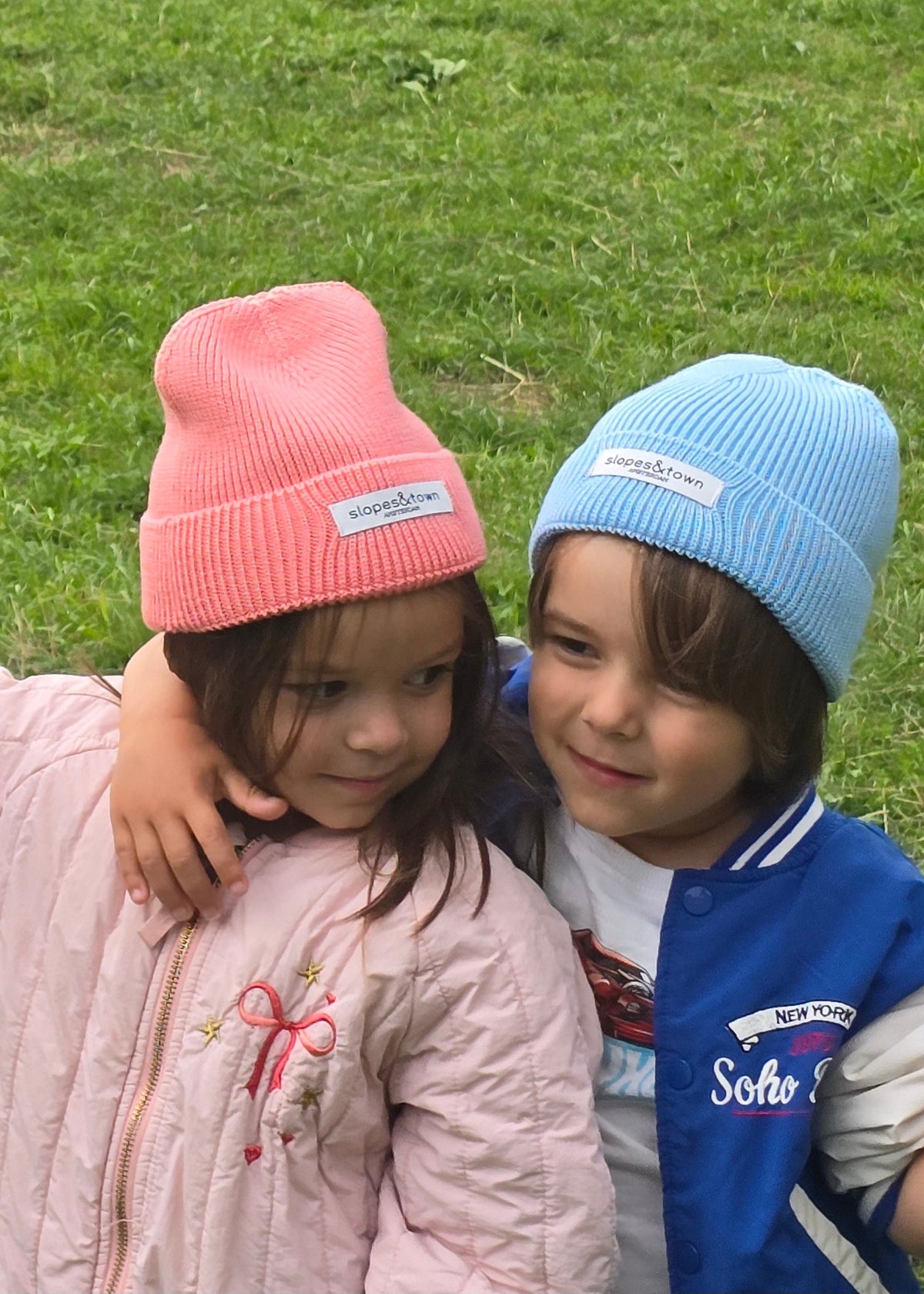 Three children standing together on a grassy lawn, each wearing a different knit beanie in yellow, pink, and light blue. The child in the center wears a pink quilted jacket, while the child on the left wears a cream cardigan and the child on the right wears a blue and white varsity jacket.
