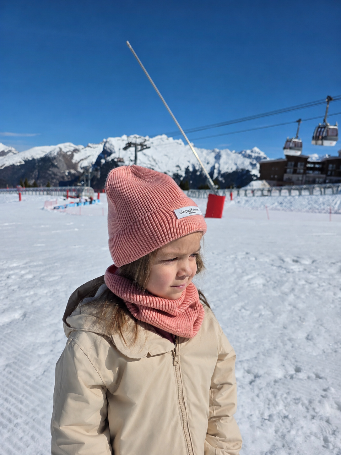A young child standing on a snowy mountain slope wearing a pink ribbed knit beanie and matching pink neck warmer, with a beige winter jacket. Ski lift cables and towers are visible in the background along with snow-covered peaks and a clear blue sky.
