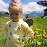 A young child wearing a pale yellow long-sleeve shirt with small blue butterfly motifs, standing outdoors in a grassy field with yellow flowers and mountains in the background.