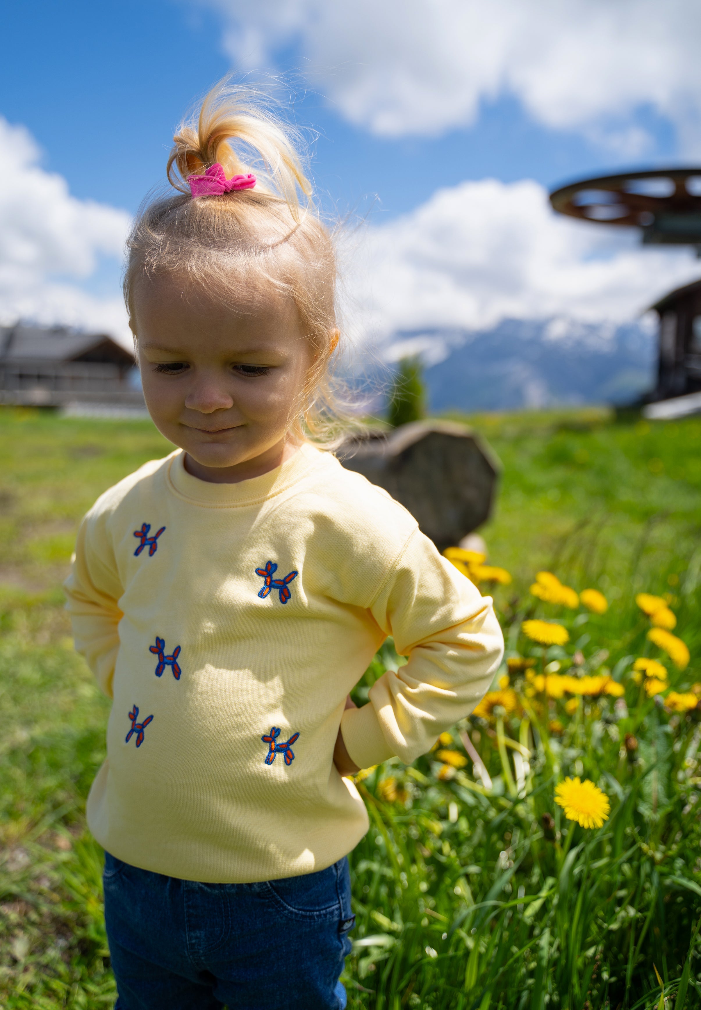 A young child wearing a pale yellow long-sleeve shirt with small blue butterfly motifs, standing outdoors in a grassy field with yellow flowers and mountains in the background.