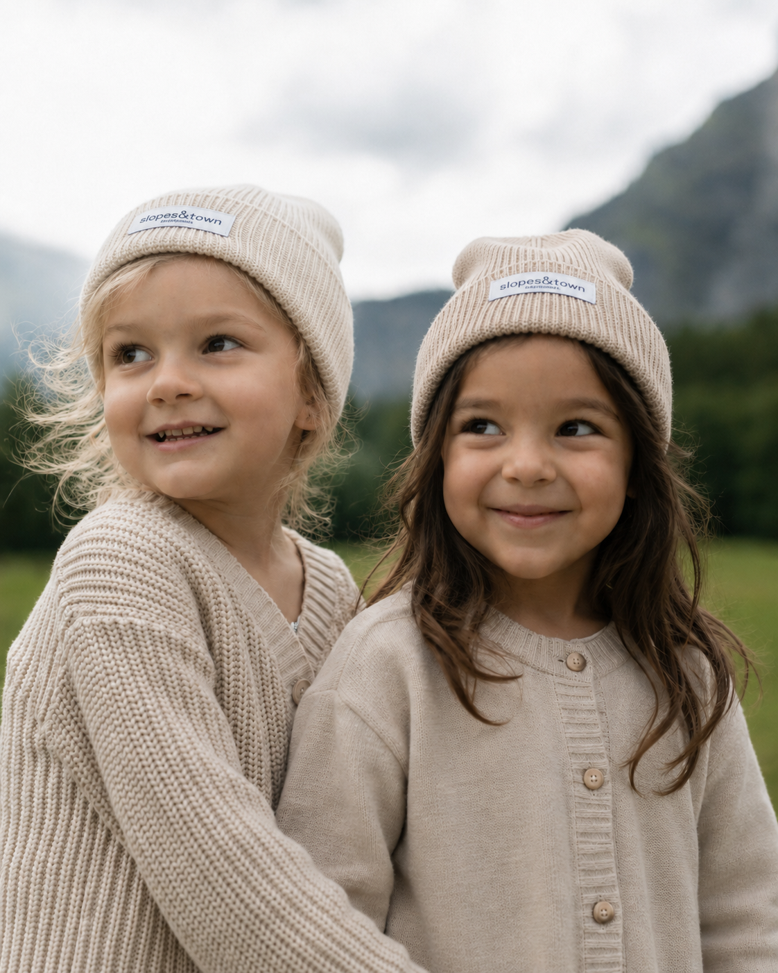 Two young children wearing matching beige knit beanie hats and coordinated cardigans, standing outdoors in front of a soft-focus mountain landscape.