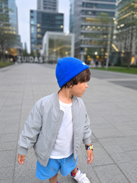 A young boy wearing a bright blue knit beanie, a light gray bomber jacket, a white t-shirt, and blue shorts, walking outdoors in a modern city plaza with blurred skyscrapers in the background.