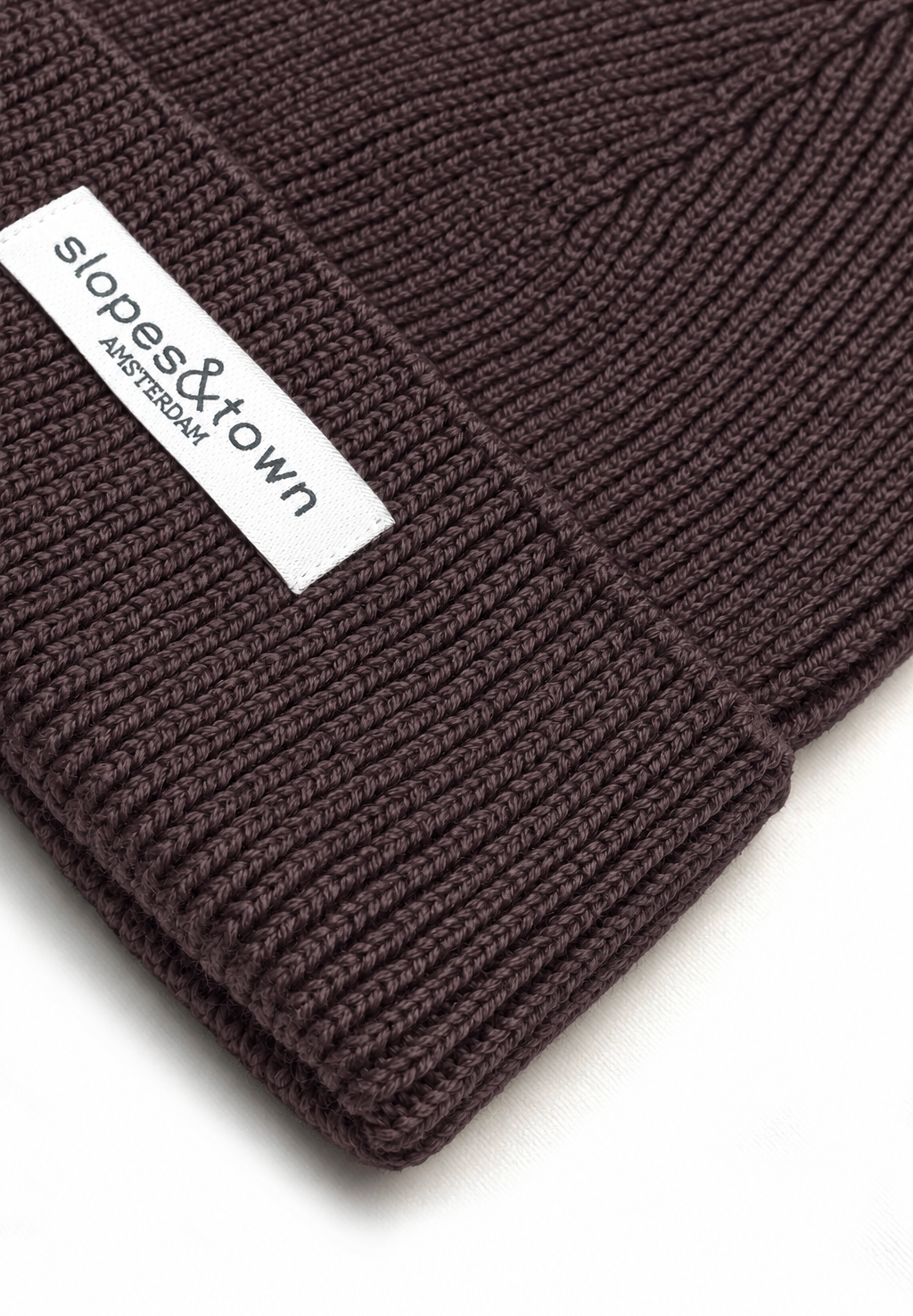 Close-up of a dark brown ribbed knit beanie with a white fabric label reading 'slopes&town AMSTERDAM' on a white background.