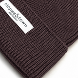 Close-up of a dark brown ribbed knit beanie with a white fabric label reading 'slopes&town AMSTERDAM' on a white background.