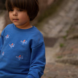 A young child wearing a bright blue crewneck sweatshirt with small red, white, and blue airplane graphics, paired with beige pants, standing outdoors on stone steps.
