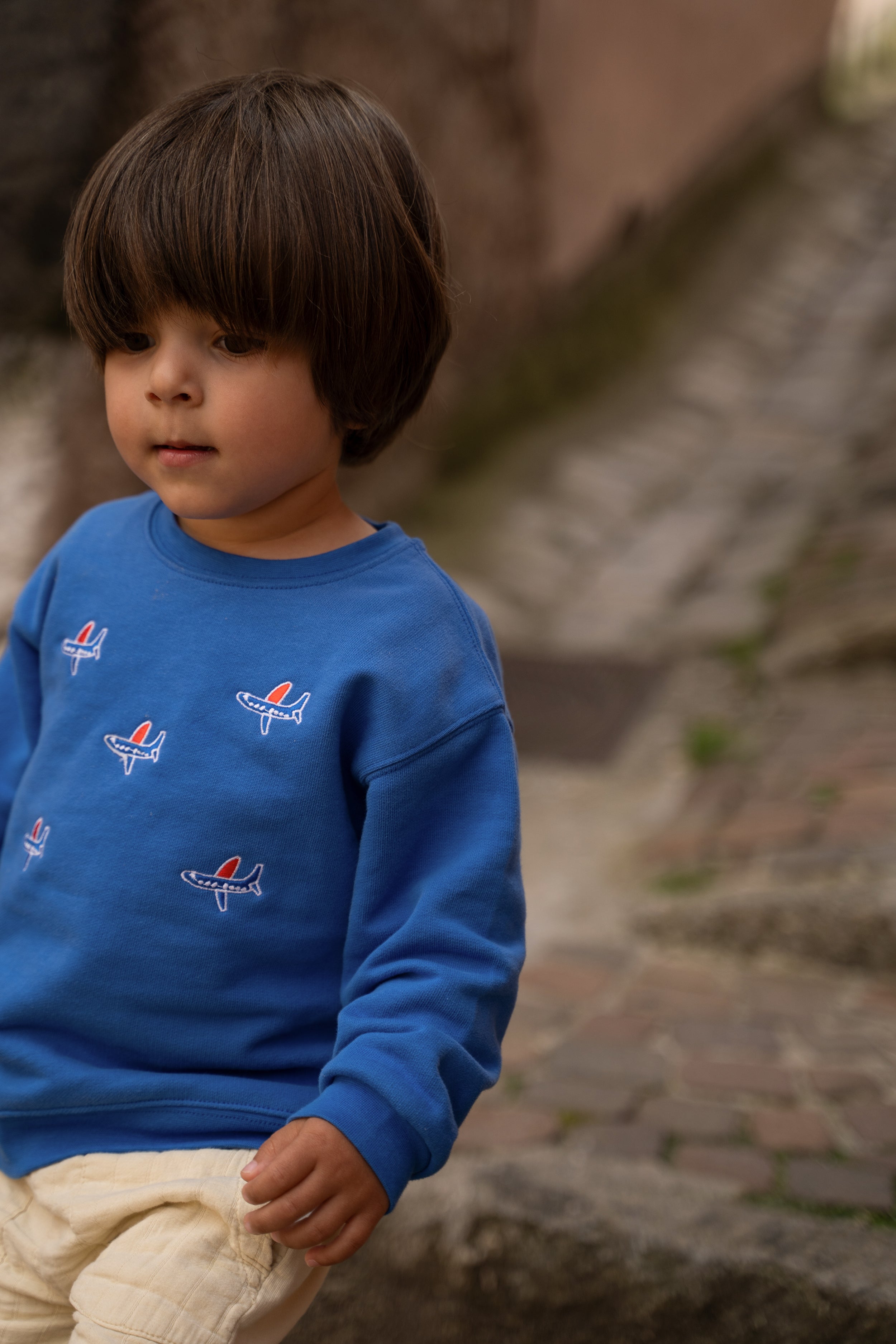 A young child wearing a bright blue crewneck sweatshirt with small red, white, and blue airplane graphics, paired with beige pants, standing outdoors on stone steps.