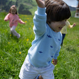 A young child wearing a light blue long-sleeve shirt with small dark blue shark prints and white shorts, running through a green grassy mountain meadow with other children blurred in the background.