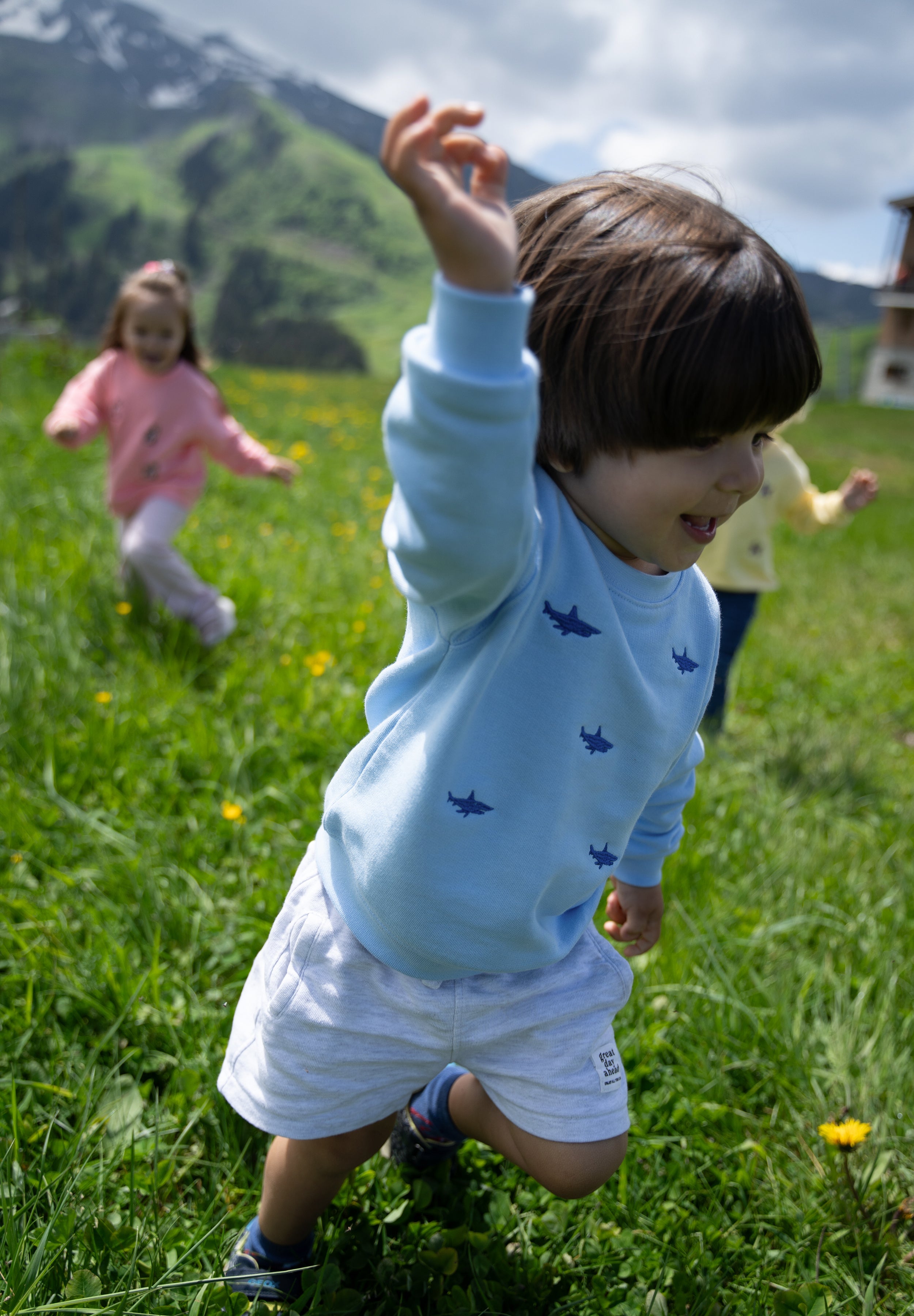 A young child wearing a light blue long-sleeve shirt with small dark blue shark prints and white shorts, running through a green grassy mountain meadow with other children blurred in the background.